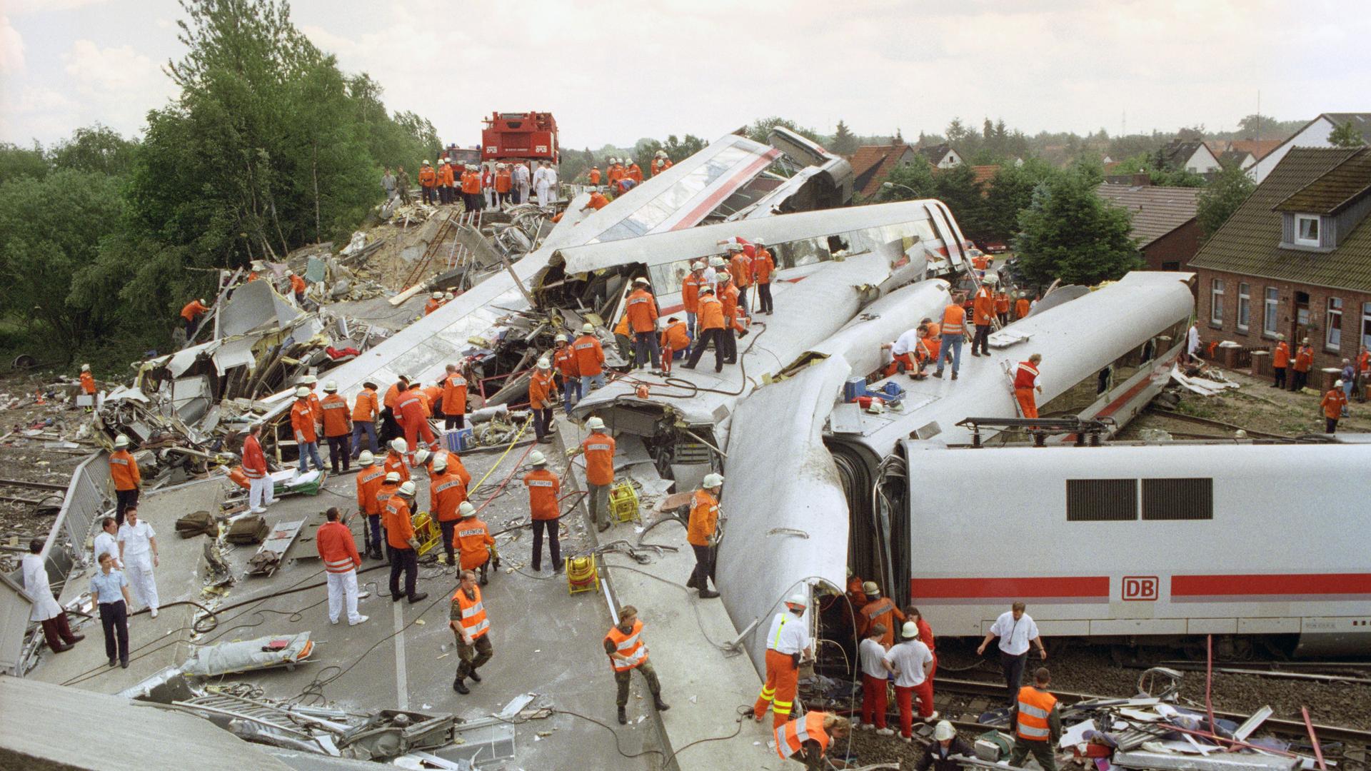 Zerstörte und ineinander verkeilte ICE-Waggons liegen am Unglücksort bei Eschede. Helfer stehen in roten Jacken auf dem Trümmerfeld. Zerstörte und ineinander verkeilte ICE-Waggons liegen am Unglücksort bei Eschede. Helfer stehen in roten Jacken auf dem Trümmerfeld.