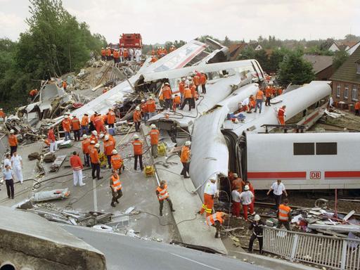 Zerstörte und ineinander verkeilte ICE-Waggons liegen am Unglücksort bei Eschede. Helfer stehen in roten Jacken auf dem Trümmerfeld.