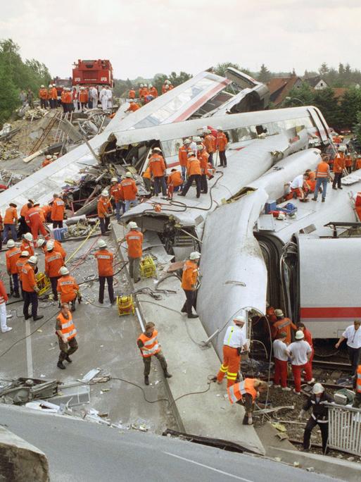 Zerstörte und ineinander verkeilte ICE-Waggons liegen am Unglücksort bei Eschede. Helfer stehen in roten Jacken auf dem Trümmerfeld.