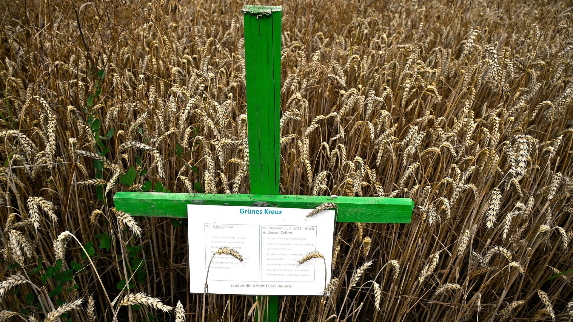 Ein grünes Kreuz in einem Weizenfeld. An dem Kreuz hängt ein Zettel, auf dem auf die Situation deutscher Landwirte hingewiesen wird. 