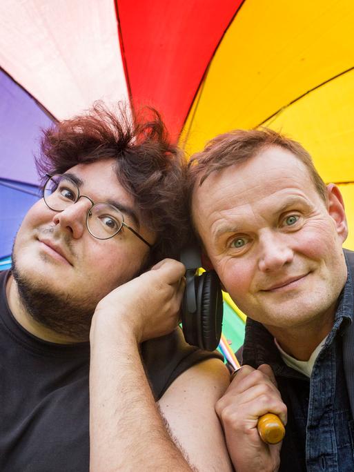 Devid Striesow und Axel Ranisch unter einem regenbogenfarbenen Regenschirm. Beide schauen freundlich in die Kamera.