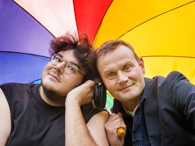 Devid Striesow und Axel Ranisch unter einem regenbogenfarbenen Regenschirm. Beide schauen freundlich in die Kamera.