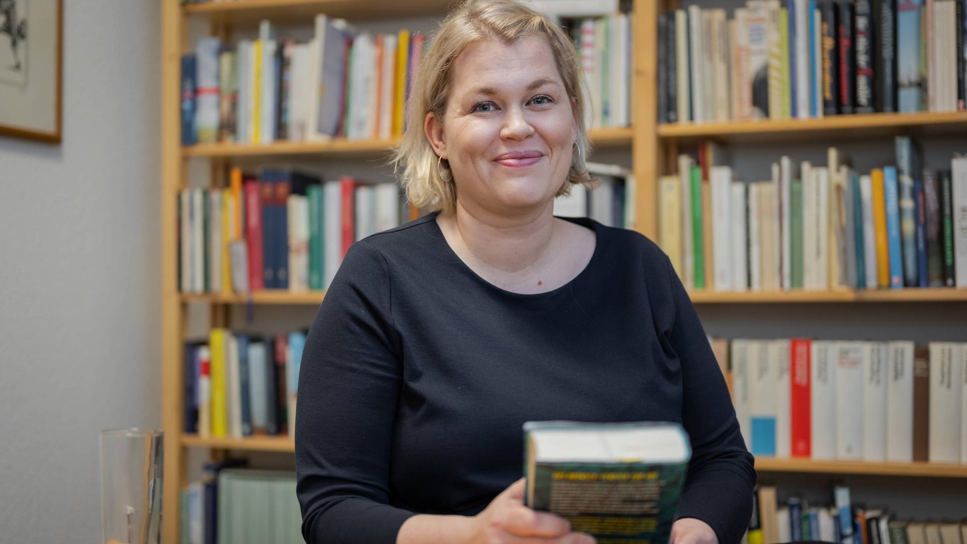 Berit Glanz steht lächelnd vor einem Bücherregal und hält selbst ein Buch in der Hand. 