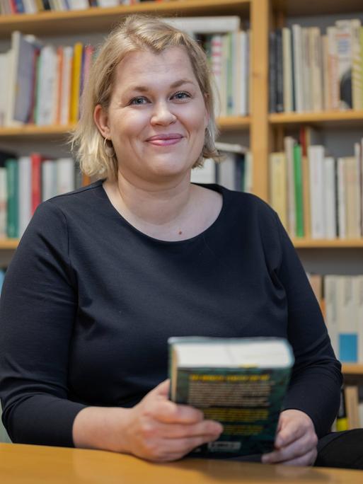 Berit Glanz steht lächelnd vor einem Bücherregal und hält selbst ein Buch in der Hand. 
