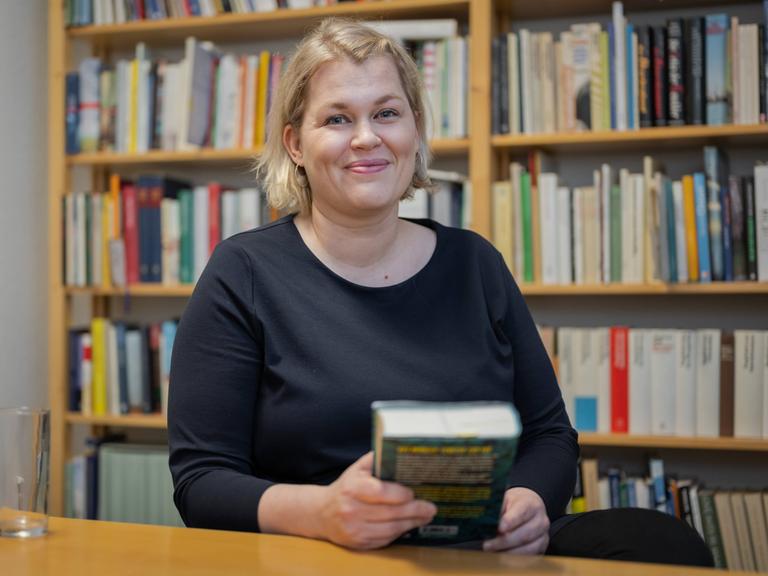 Berit Glanz steht lächelnd vor einem Bücherregal und hält selbst ein Buch in der Hand. 