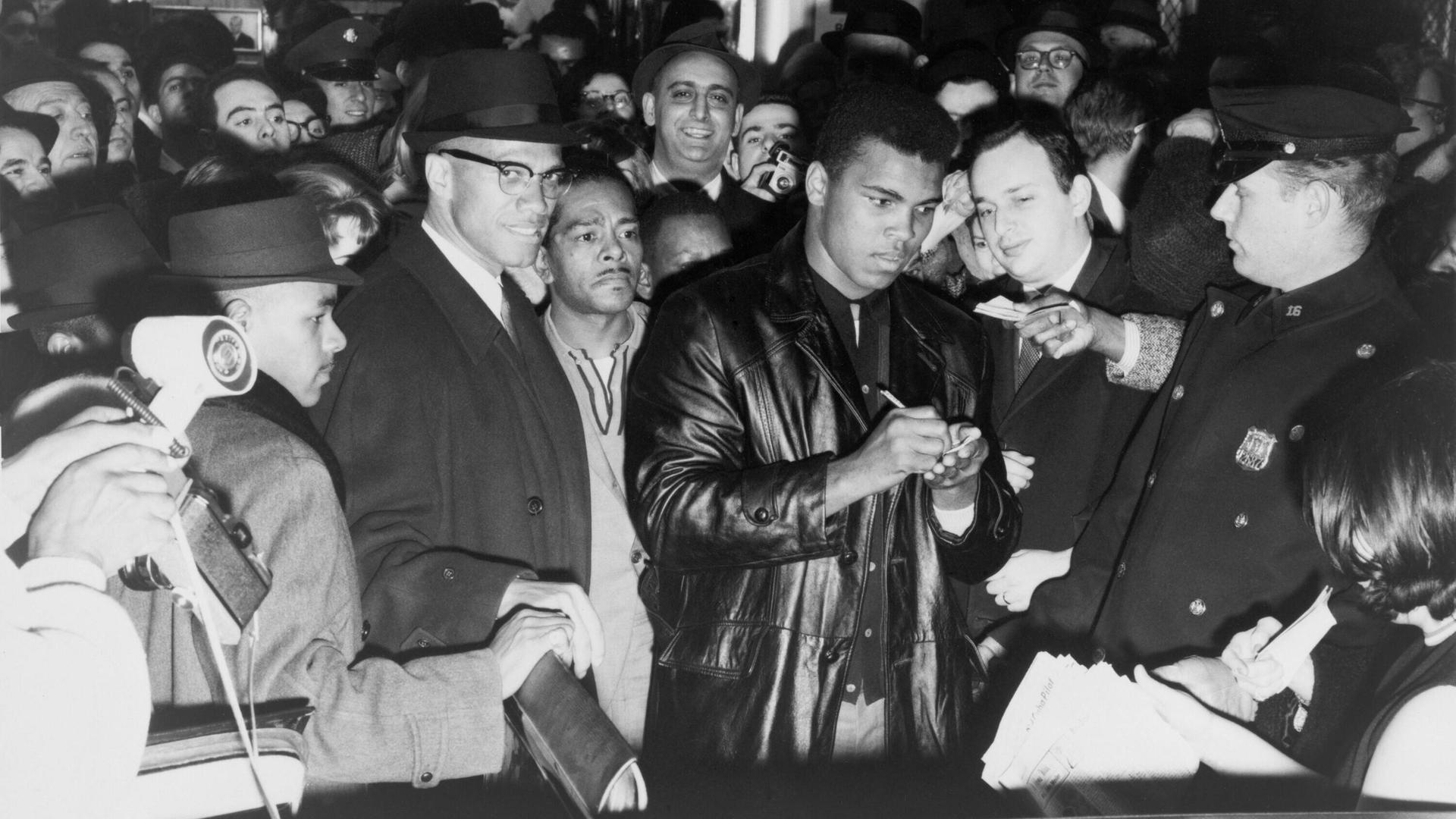 Muhammad Ali (Cassius Clay) schreibt nach dem Gewinn des Weltmeistertitels Autogramme in New York. Zu seiner Rechten steht mit Brille und Hut der politische Aktivist Malcom X.