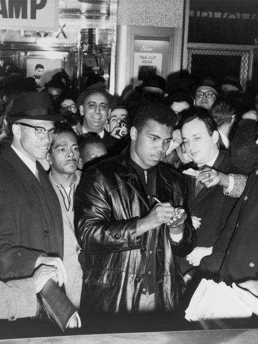 Muhammad Ali (Cassius Clay) schreibt nach dem Gewinn des Weltmeistertitels Autogramme in New York. Zu seiner Rechten steht mit Brille und Hut der politische Aktivist Malcom X.