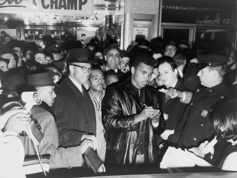 Muhammad Ali (Cassius Clay) schreibt nach dem Gewinn des Weltmeistertitels Autogramme in New York. Zu seiner Rechten steht mit Brille und Hut der politische Aktivist Malcom X.