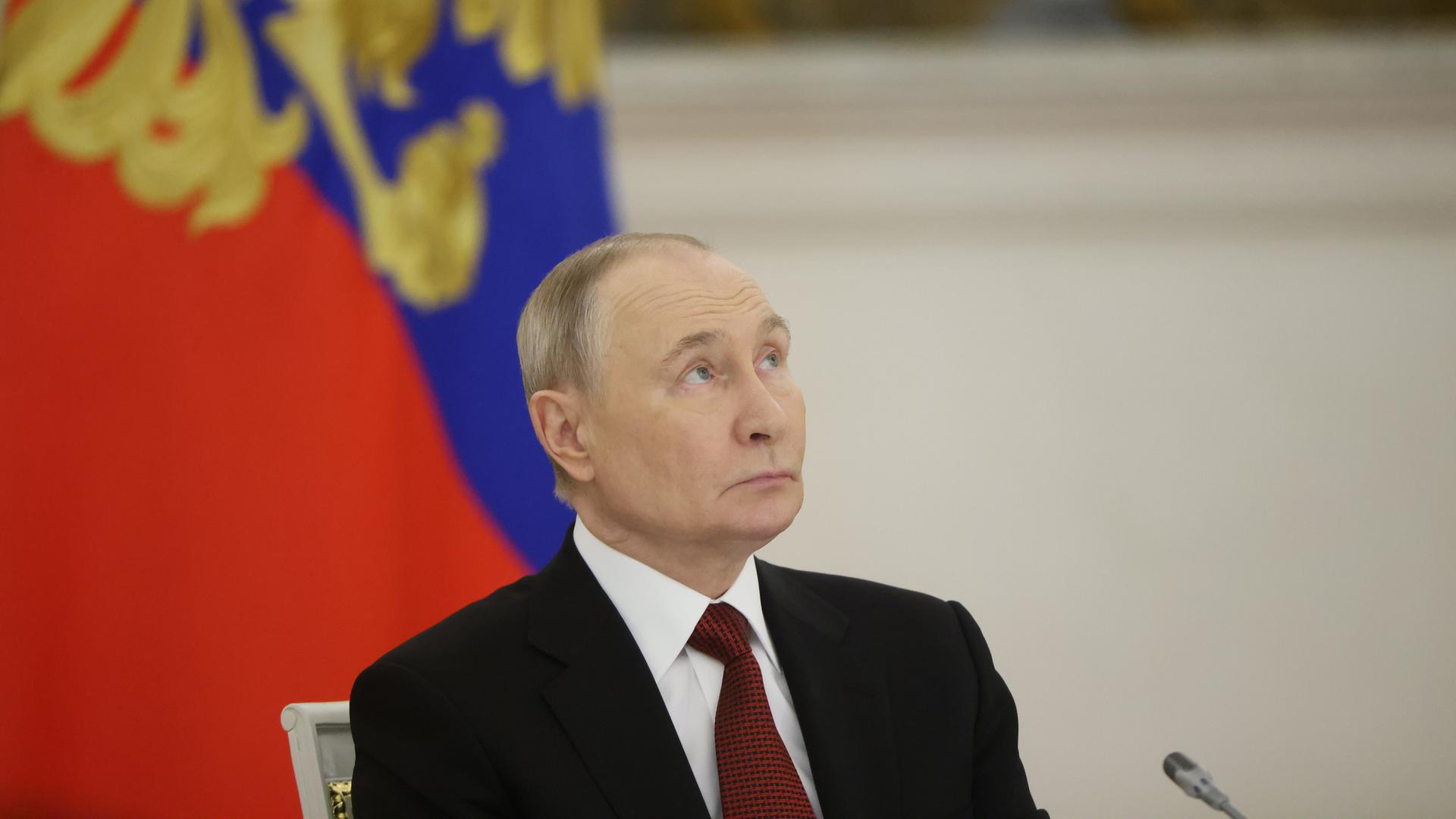 Portrait von Putin, der vor einer russischen Flagge leicht nach oben schaut.