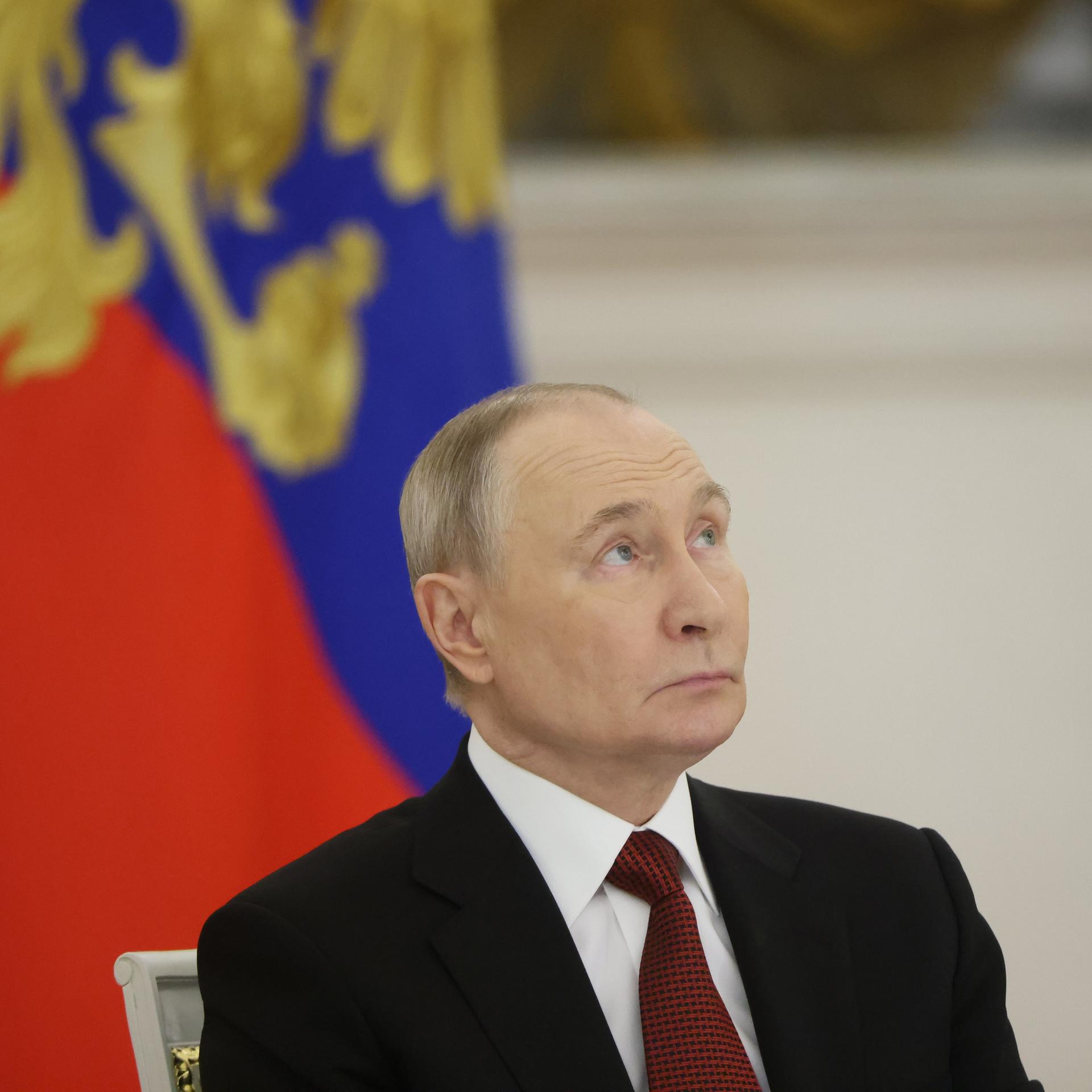 Portrait von Putin, der vor einer russischen Flagge leicht nach oben schaut.