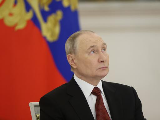 Portrait von Putin, der vor einer russischen Flagge leicht nach oben schaut.