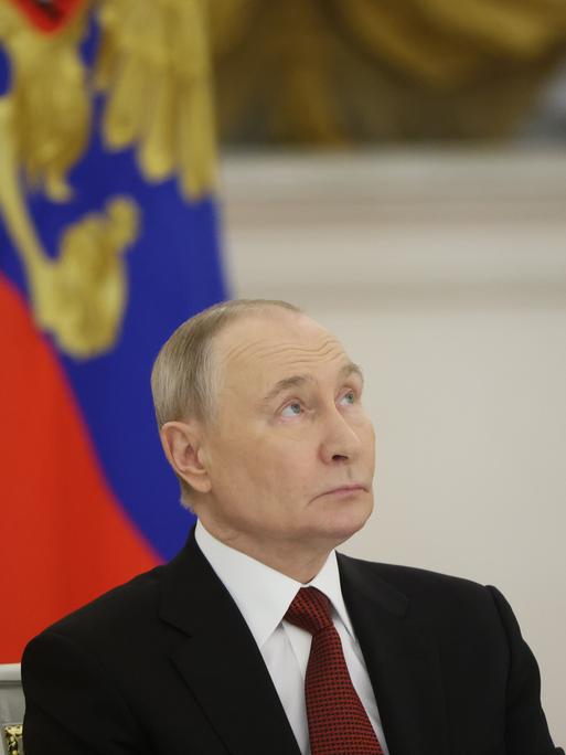 Portrait von Putin, der vor einer russischen Flagge leicht nach oben schaut.