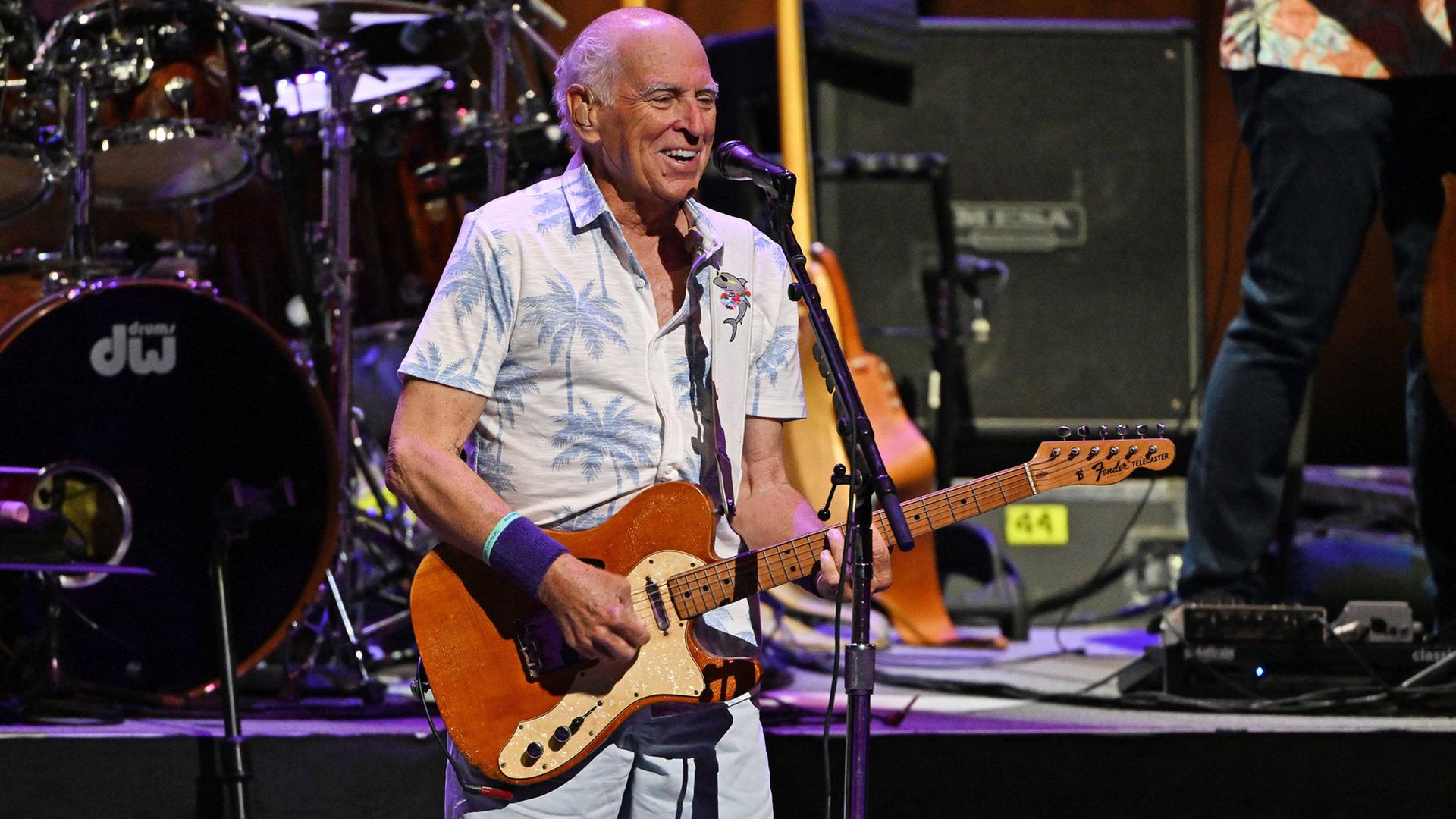 Das Foto zeigt den amerikanischen Singer-Songwriter Jimmy Buffett. Er steht mit einer Gitarre auf einer Bühne.