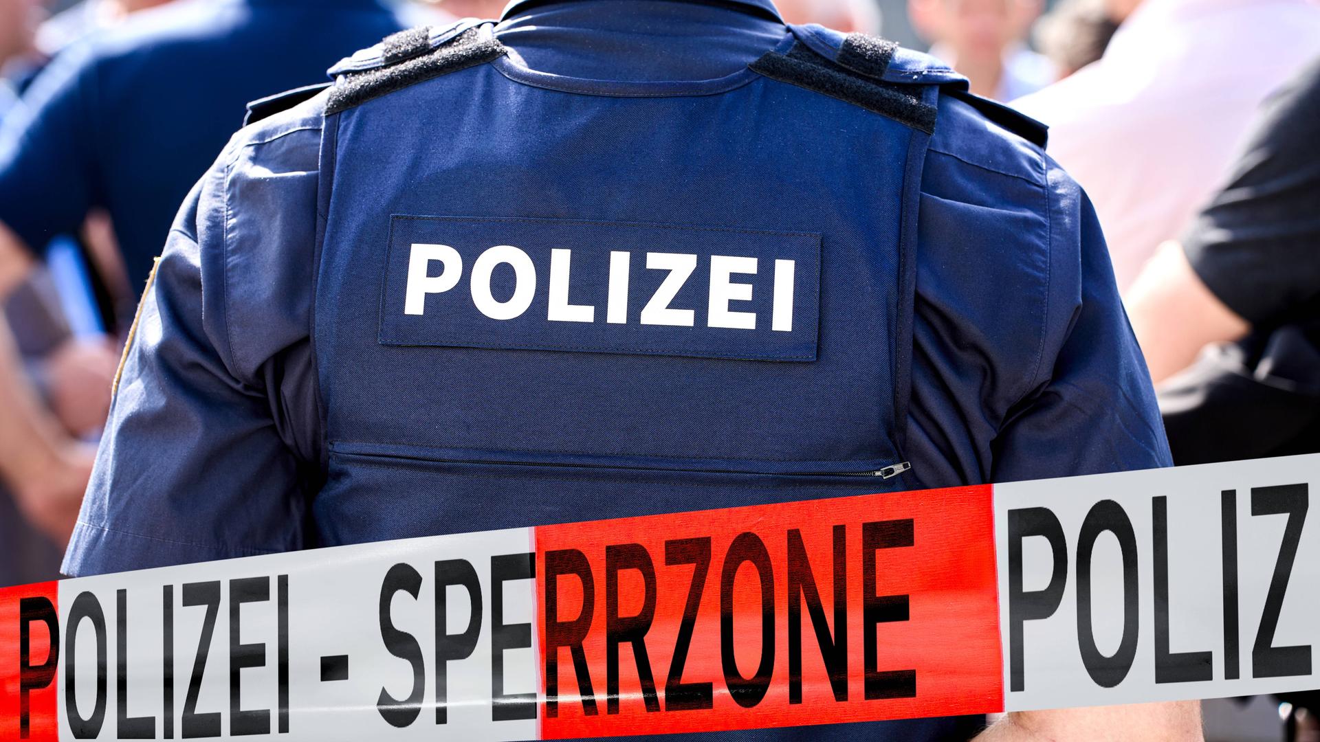 Ein Polizeibeamter von hinten in dunkelblauen Uniform mit der Aufschrift POLIZEI. Eine Polizei-Absperrung mit der Aufschrift SPERRZONE, im Hintergrund sind mehrere Personen einer Menschenansammlung.