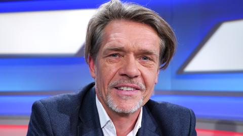 Ein Mann mittleren Alters mit grauen Haaren und modischem Haarschnitt in einem Fernsehstudio