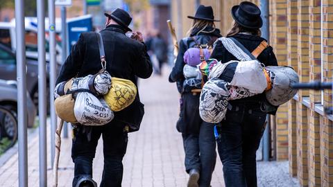 Blick von hinten auf zwei Wandergesellinnen und einen Wandergesellen, die in traditoneller Handwerker-Kluft durch eine Straße laufen.  