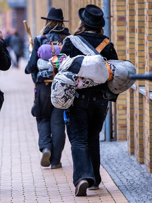 Blick von hinten auf zwei Wandergesellinnen und einen Wandergesellen, die in traditoneller Handwerker-Kluft durch eine Straße laufen.  