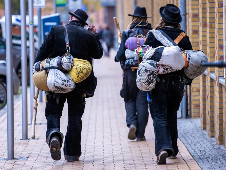 Blick von hinten auf zwei Wandergesellinnen und einen Wandergesellen, die in traditoneller Handwerker-Kluft durch eine Straße laufen.  
