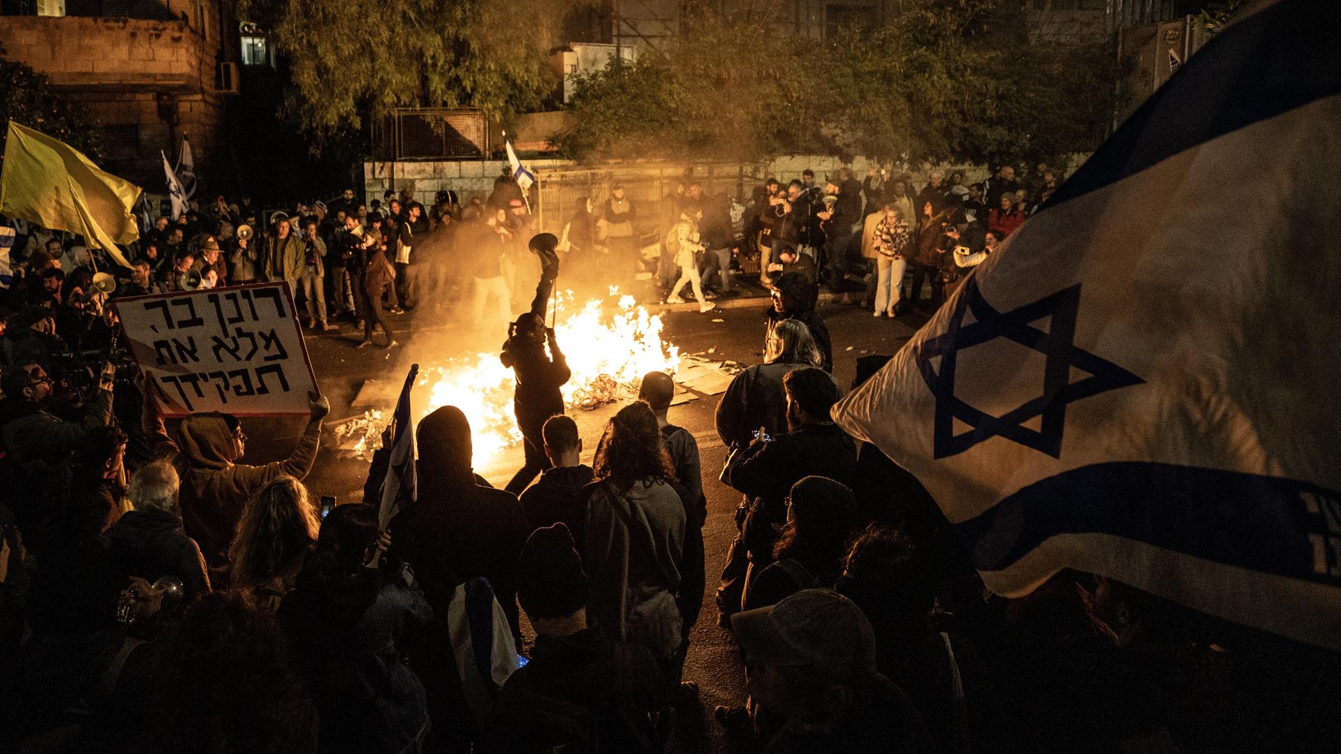 Ein Feuer erhellt die nächtliche Szene. Auf der Straße sind viele Demonstranten mit israelischen Flaggen und Transparenten mit hebräischer Aufschrift unterwegs.