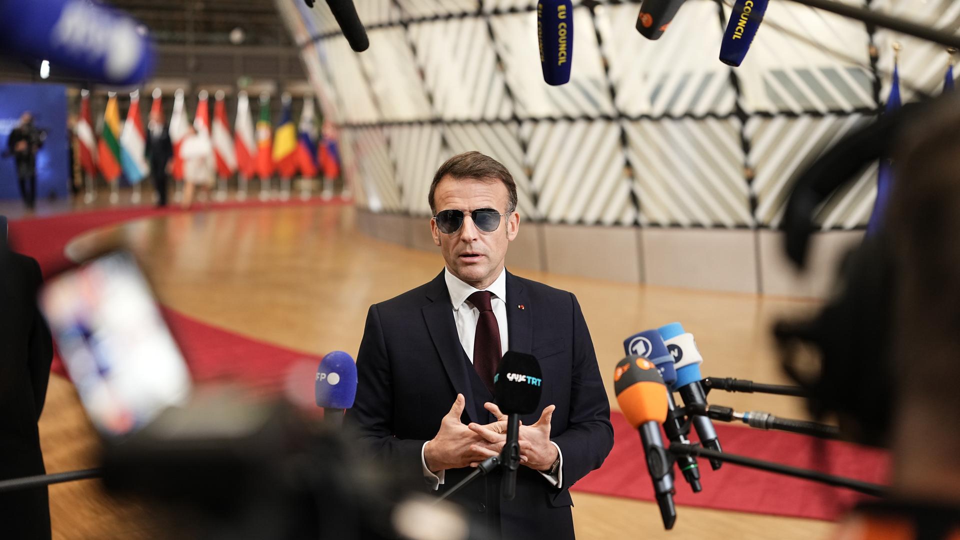 Der französische Präsident Emmanuel Macron trägt im Vorfeld eines Sondergipfels des Europäischen Rates eine Sonnenbrille und spricht in zahlreiche Mikrofone. Im Hintergrund bunte Flaggen.