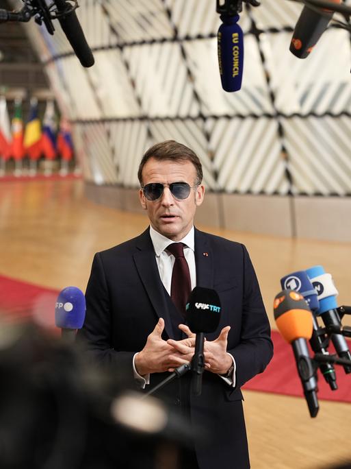 Der französische Präsident Emmanuel Macron trägt im Vorfeld eines Sondergipfels des Europäischen Rates eine Sonnenbrille und spricht in zahlreiche Mikrofone. Im Hintergrund bunte Flaggen.