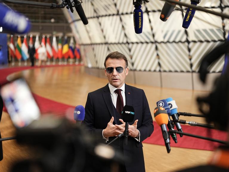 Der französische Präsident Emmanuel Macron trägt im Vorfeld eines Sondergipfels des Europäischen Rates eine Sonnenbrille und spricht in zahlreiche Mikrofone. Im Hintergrund bunte Flaggen.