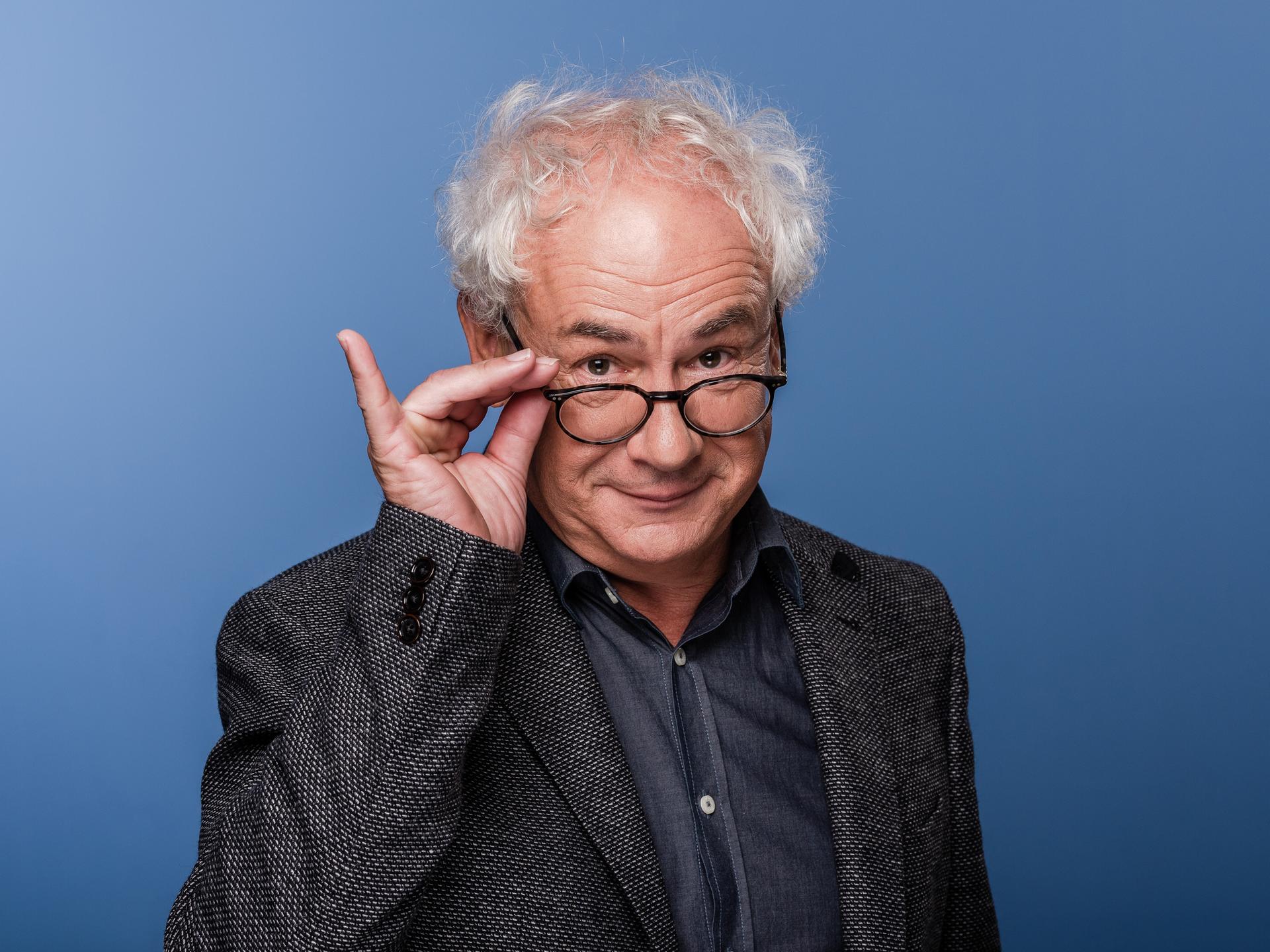 Schauspieler Tom Pauls mit der Hand an seiner Brille vor blauem Hinterund. 