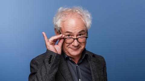 Schauspieler Tom Pauls mit der Hand an seiner Brille vor blauem Hinterund. Schauspieler Tom Pauls mit der Hand an seiner Brille vor blauem Hinterund.