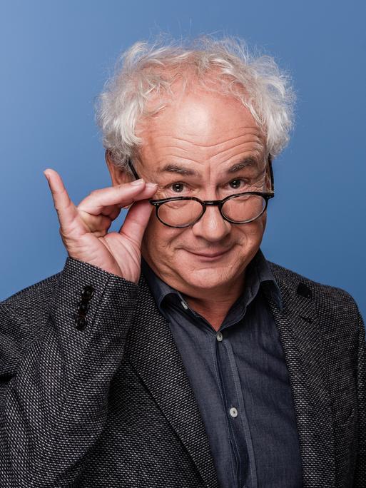 Schauspieler Tom Pauls mit der Hand an seiner Brille vor blauem Hinterund. 