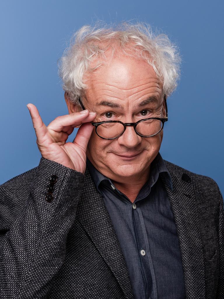 Schauspieler Tom Pauls mit der Hand an seiner Brille vor blauem Hinterund. 