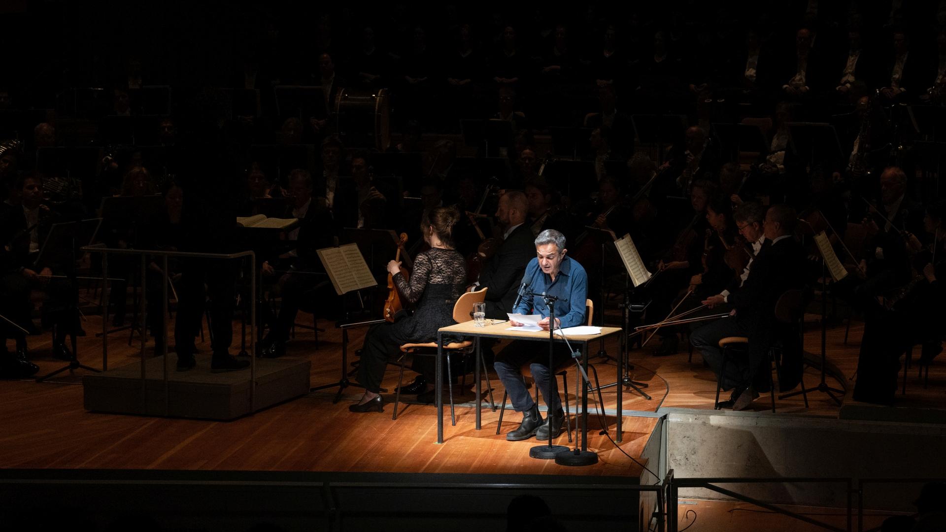 Navid Kermani sitzt auf einer Bühne inmitten eines Lichtkegels, um ihn herum kann man die Musiker des Orchesters erahnen.