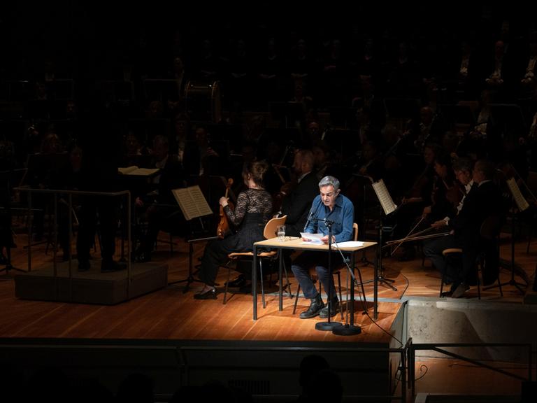 Navid Kermani sitzt auf einer Bühne inmitten eines Lichtkegels, um ihn herum kann man die Musiker des Orchesters erahnen.