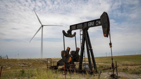 Eine kleine Ölförderpumpe steht auf einem Feld, dahinter steht eine Windturbine, am Horizont viele weitere. Eine kleine Ölförderpumpe steht auf einem Feld, dahinter steht eine Windturbine, am Horizont viele weitere.