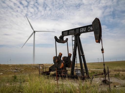 Eine kleine Ölförderpumpe steht auf einem Feld, dahinter steht eine Windturbine, am Horizont viele weitere.