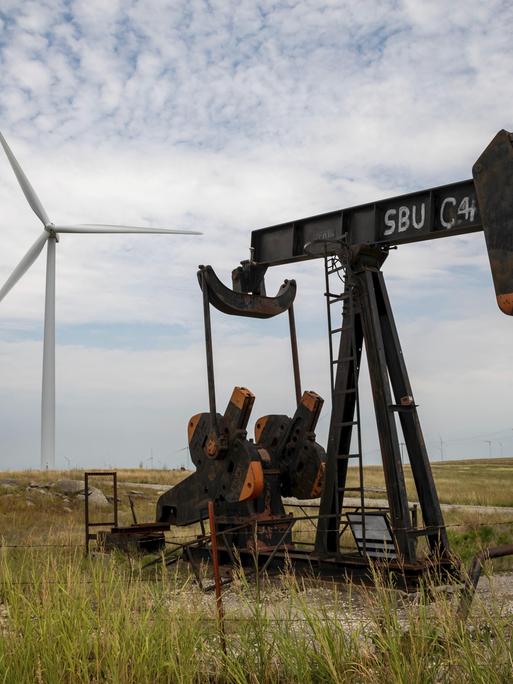 Eine kleine Ölförderpumpe steht auf einem Feld, dahinter steht eine Windturbine, am Horizont viele weitere.