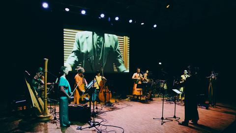Musiker und Musikerinnen, unter anderem mit Harfe und Kontrabass stehen auf einer bunt beleuchteten Bühne, auf der im Hintergrund ein Screen hängt, der gerade einen Anzugträger zeigt.