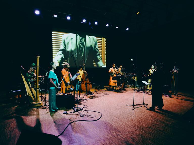 Musiker und Musikerinnen, unter anderem mit Harfe und Kontrabass stehen auf einer bunt beleuchteten Bühne, auf der im Hintergrund ein Screen hängt, der gerade einen Anzugträger zeigt.