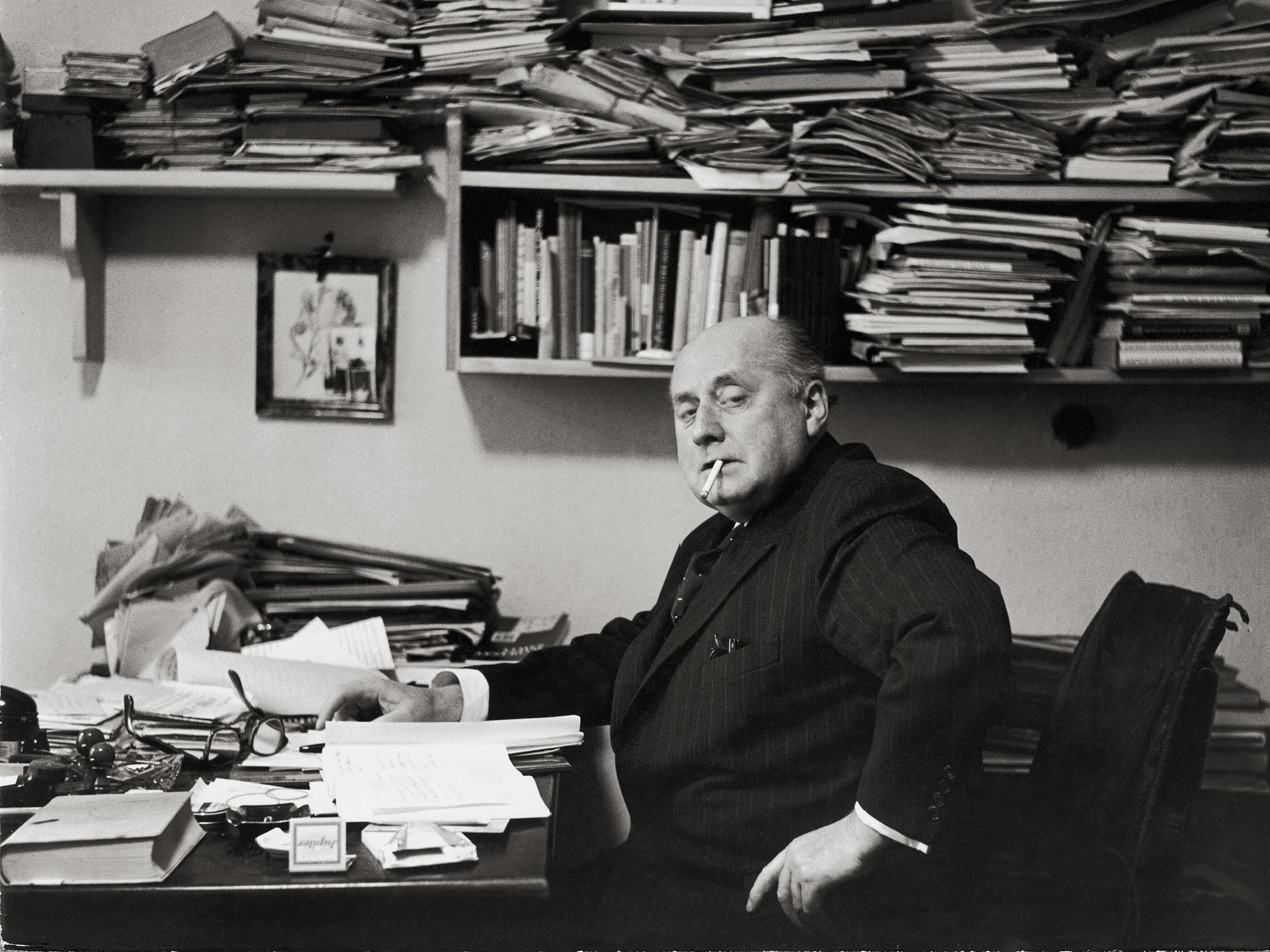 Gottfried Benn in seiner Berliner Wohnung. Regale und Tisch sind überfüllt mit Büchern und Notizen. Benn raucht.