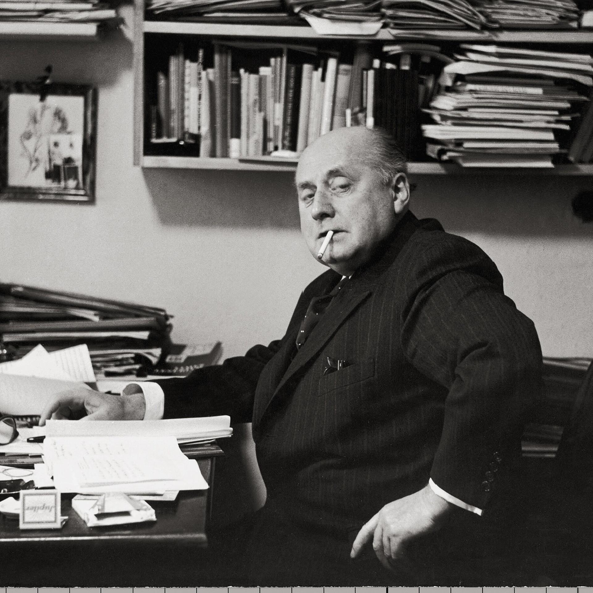 Gottfried Benn in seiner Berliner Wohnung. Regale und Tisch sind überfüllt mit Büchern und Notizen. Benn raucht.