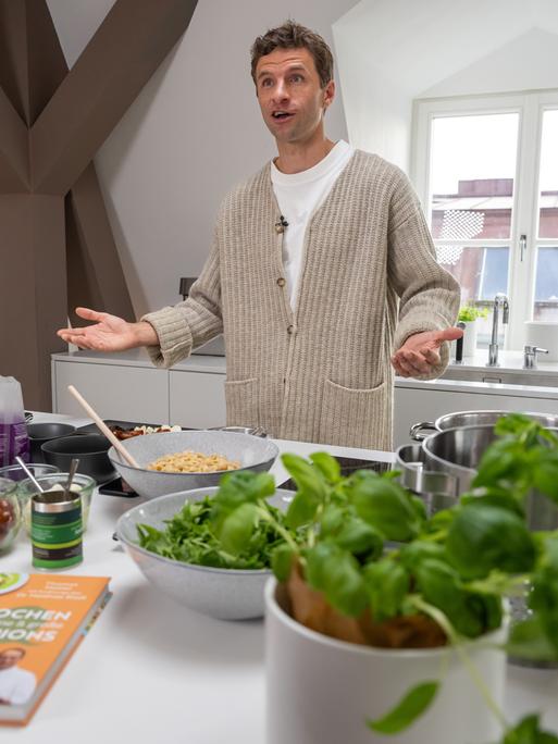 Ein Mann steht mit ausgebreiteten Händen in einer Küche. Vor ihm liegen verschiedene, überwiegend frische Zutaten und ein Buch.
