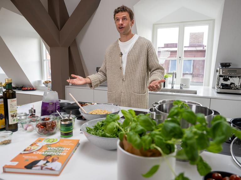 Ein Mann steht mit ausgebreiteten Händen in einer Küche. Vor ihm liegen verschiedene, überwiegend frische Zutaten und ein Buch.