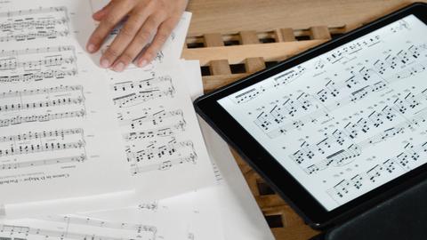 Eine Hand bewegt sich auf einem Tisch, auf dem ausgedruckte Notenblätter und ein Tablet mit Noten liegen.