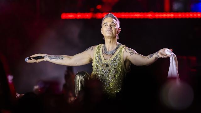 Robbie Williams steht auf einer Bühne und breitet die Arme aus.