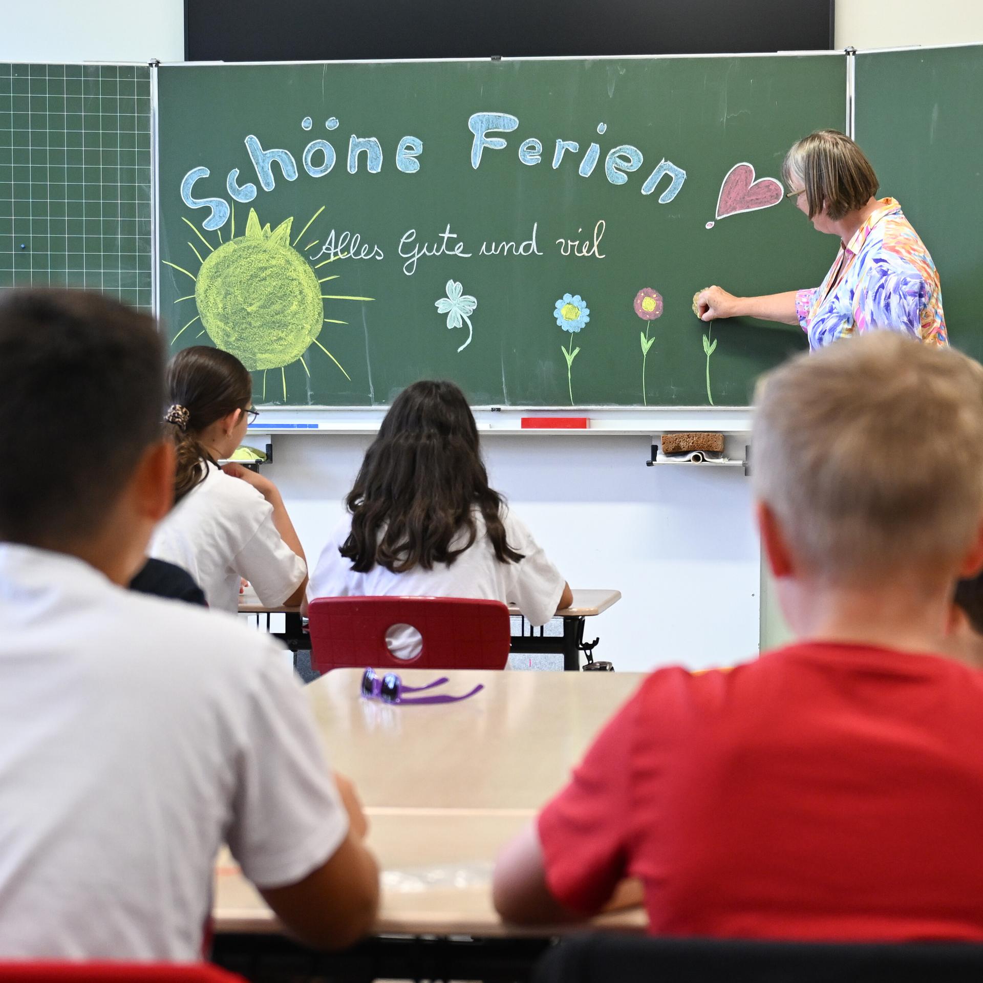 Schulkinder sitzen im Klassenzimmer am letzten Schultag vor den Sommerferien, eine Lehrerin schreibt an eine Tafel "Schöne Ferien Alles Gute und viel Glück".
