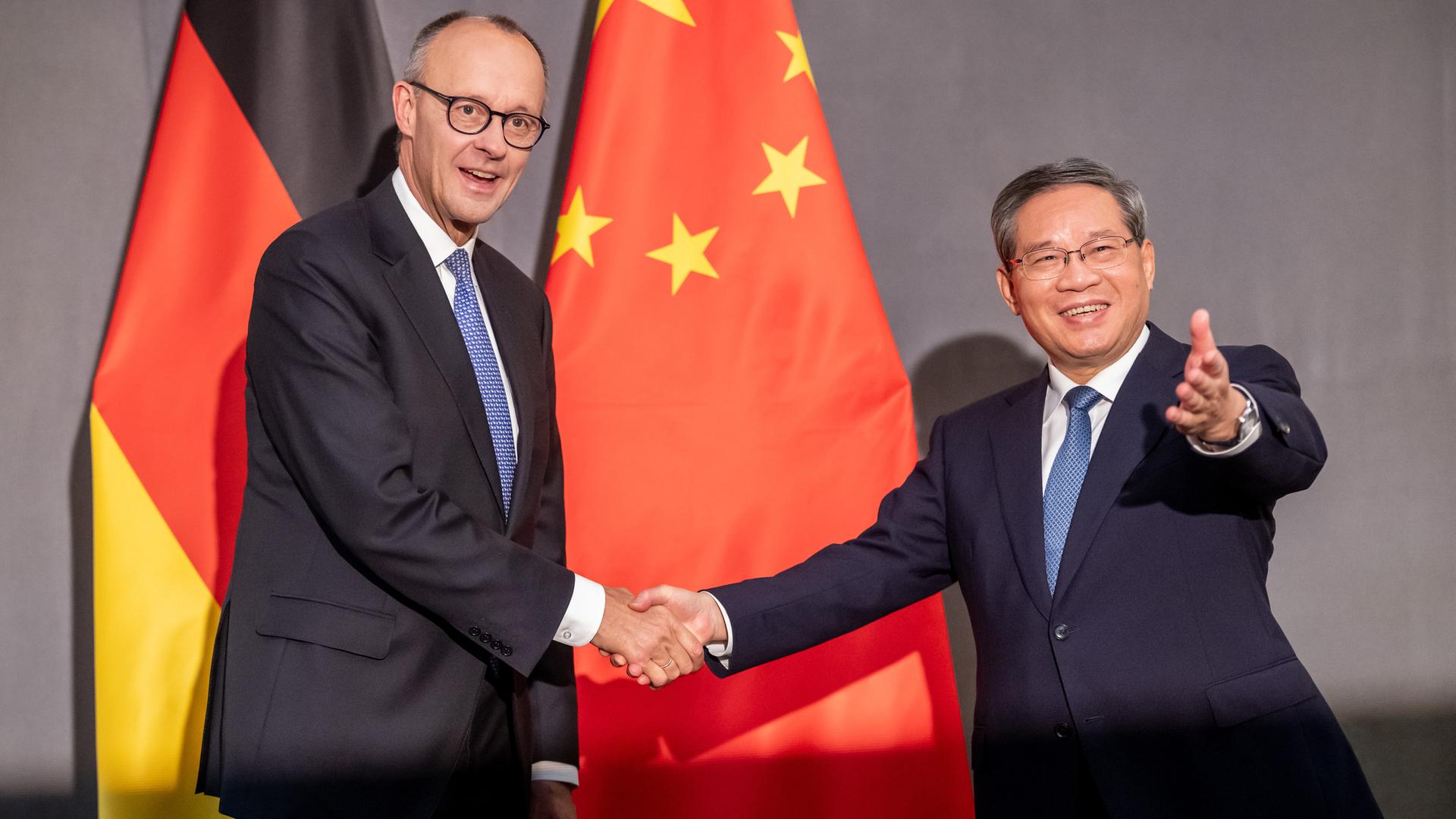 Friedrich Merz und Li Qiang reichen sich lächelnd die Hand. Im Hintergrund sind eine deutsche und eine chinesische Flagge zu sehen.