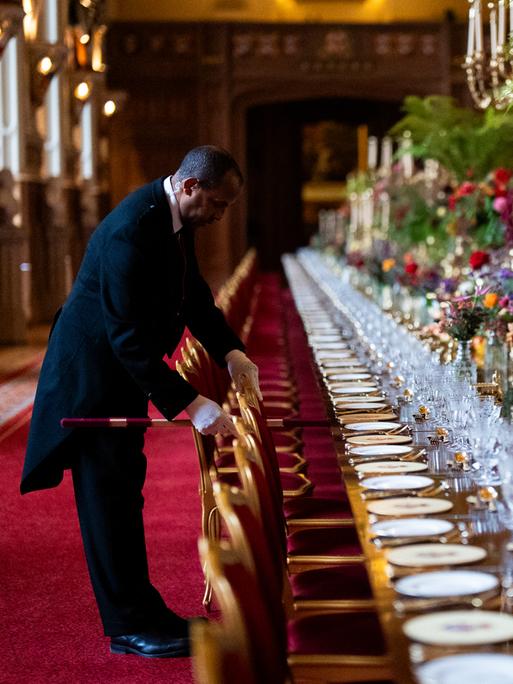 Die Stühle am elegant gedecktem Banketttisch in der St. Georges Hall werden von einem Angestellten im richtigen Abstand zueinander aufgestellt im Vorfeld eines Staatsbanketts im Windsor Castle in England. 