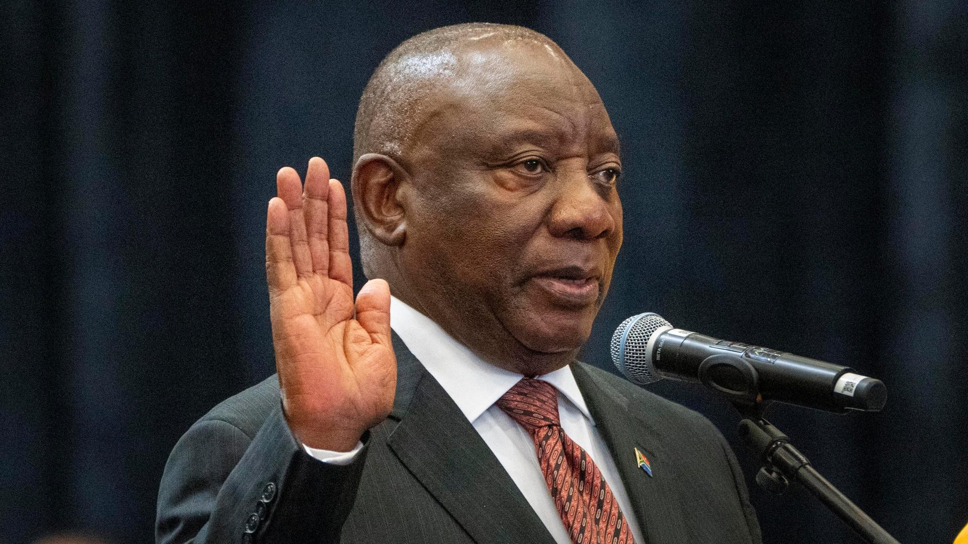 Cyril Ramaphosa steht an einem Mikrofon und hebt die Hand.