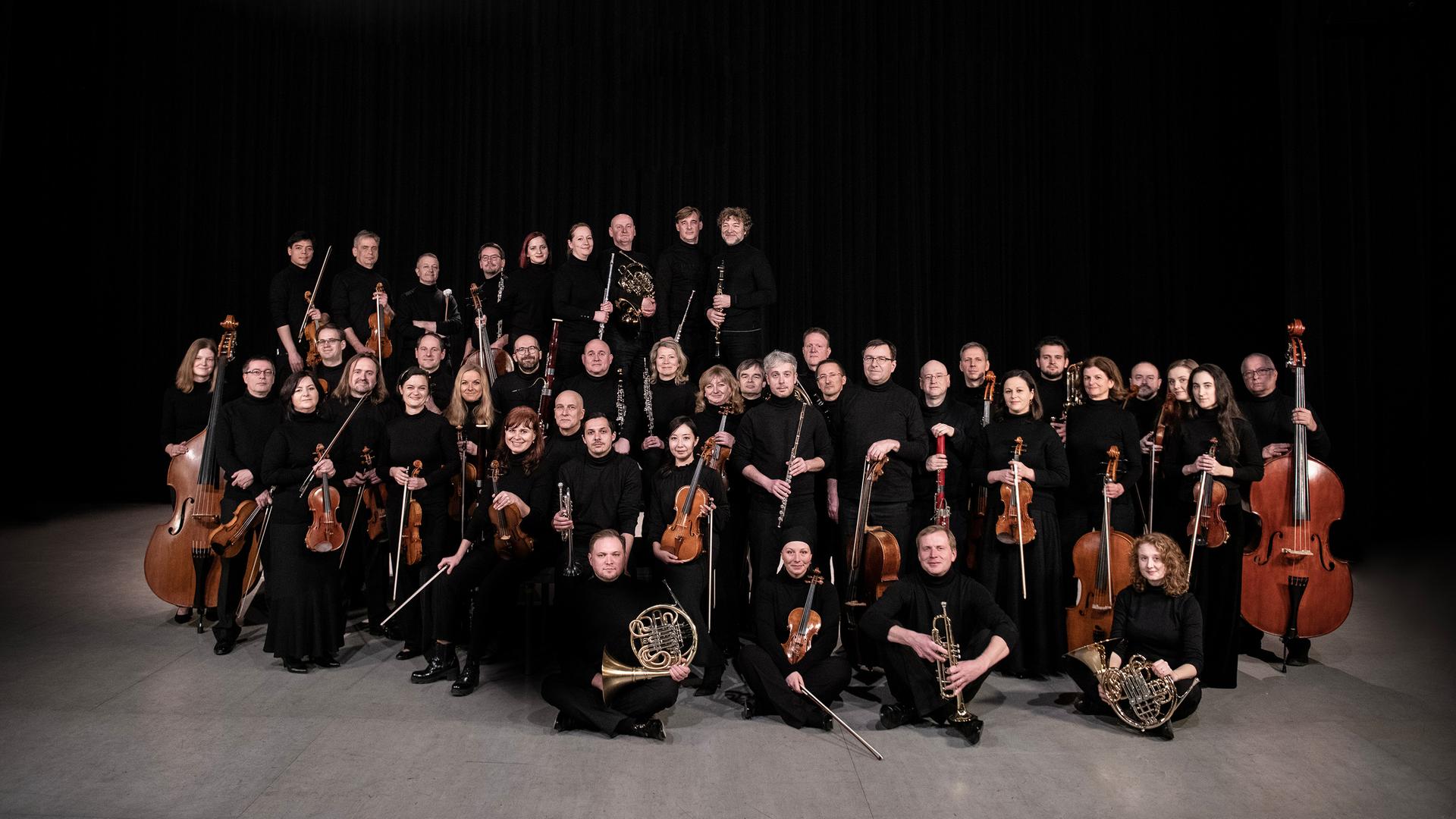 Das Orchester steht und sitzt in einer wolkenartigen Formation beieinander. Alle haben ihre Instrumente dabei. Das Orchester steht und sitzt in einer wolkenartigen Formation beieinander. Alle haben ihre Instrumente dabei.