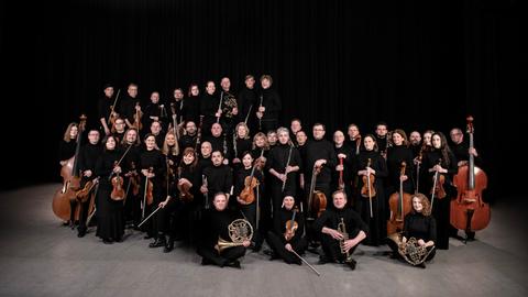 Das Orchester steht und sitzt in einer wolkenartigen Formation beieinander. Alle haben ihre Instrumente dabei.