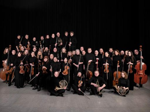 Das Orchester steht und sitzt in einer wolkenartigen Formation beieinander. Alle haben ihre Instrumente dabei.